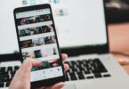 Person browsing social media on a phone in front of a laptop, switching to Instagram on PC