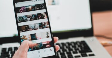Person browsing social media on a phone in front of a laptop, switching to Instagram on PC