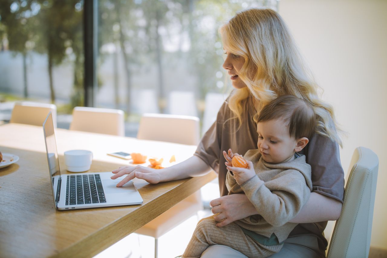 Parent holding a baby while working on a laptop to make money from home