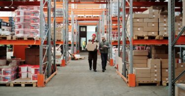 Warehouse workers carrying a package past stocked shelves, illustrating a Wholesale2b dropshipping supplier