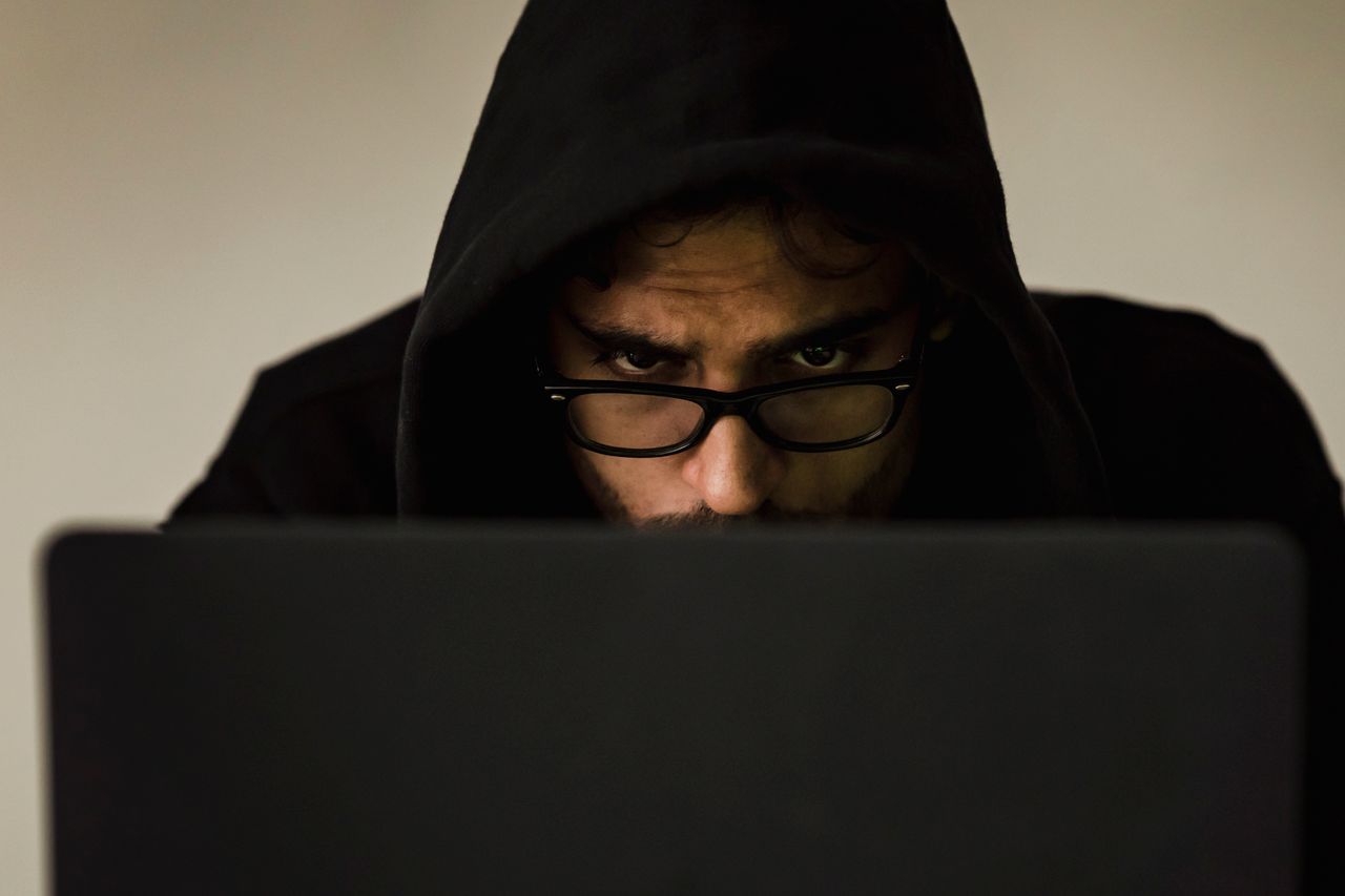 Hooded scammer using a laptop, the kind of operator behind fake online earning sites