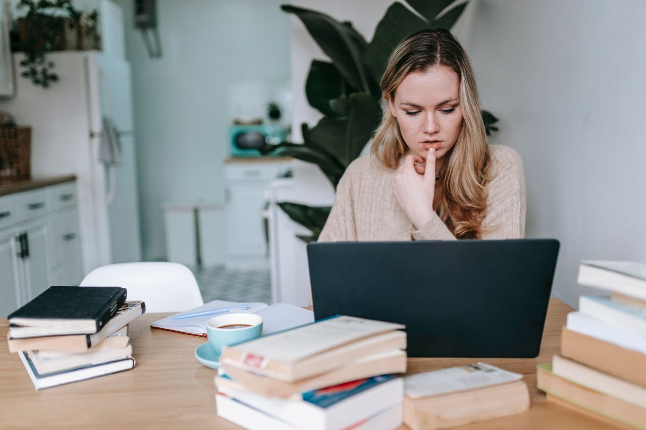 Thoughtful student using a laptop at a table of books while comparing online jobs for students