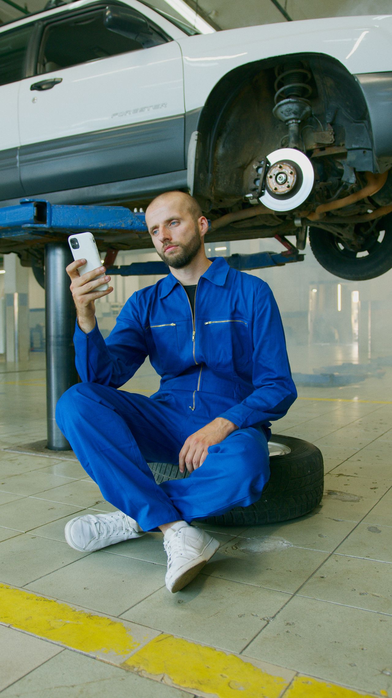 Mechanic sitting on a tire using a smartphone, weighing X-Cart alternatives for an auto parts store