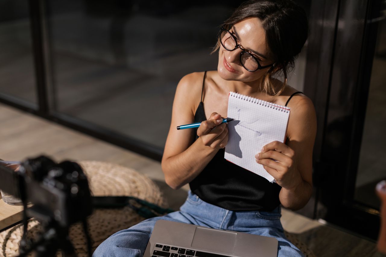 Smiling creator vlogging with a camera, laptop and notepad, the kind of seller Podia targets